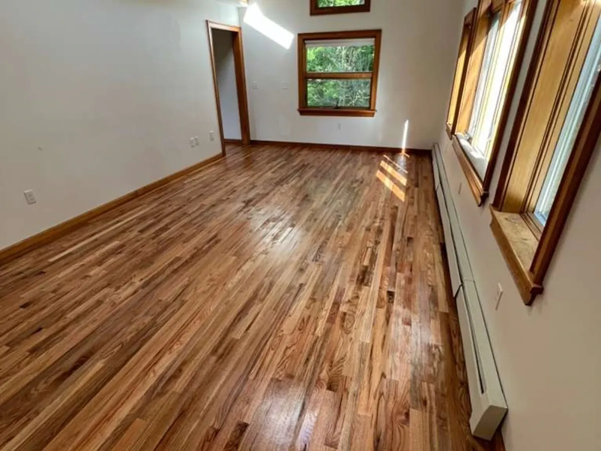 Glossy hardwood flooring in a bright room, with visible grain variation, wood baseboards, and tall wood-trim windows.