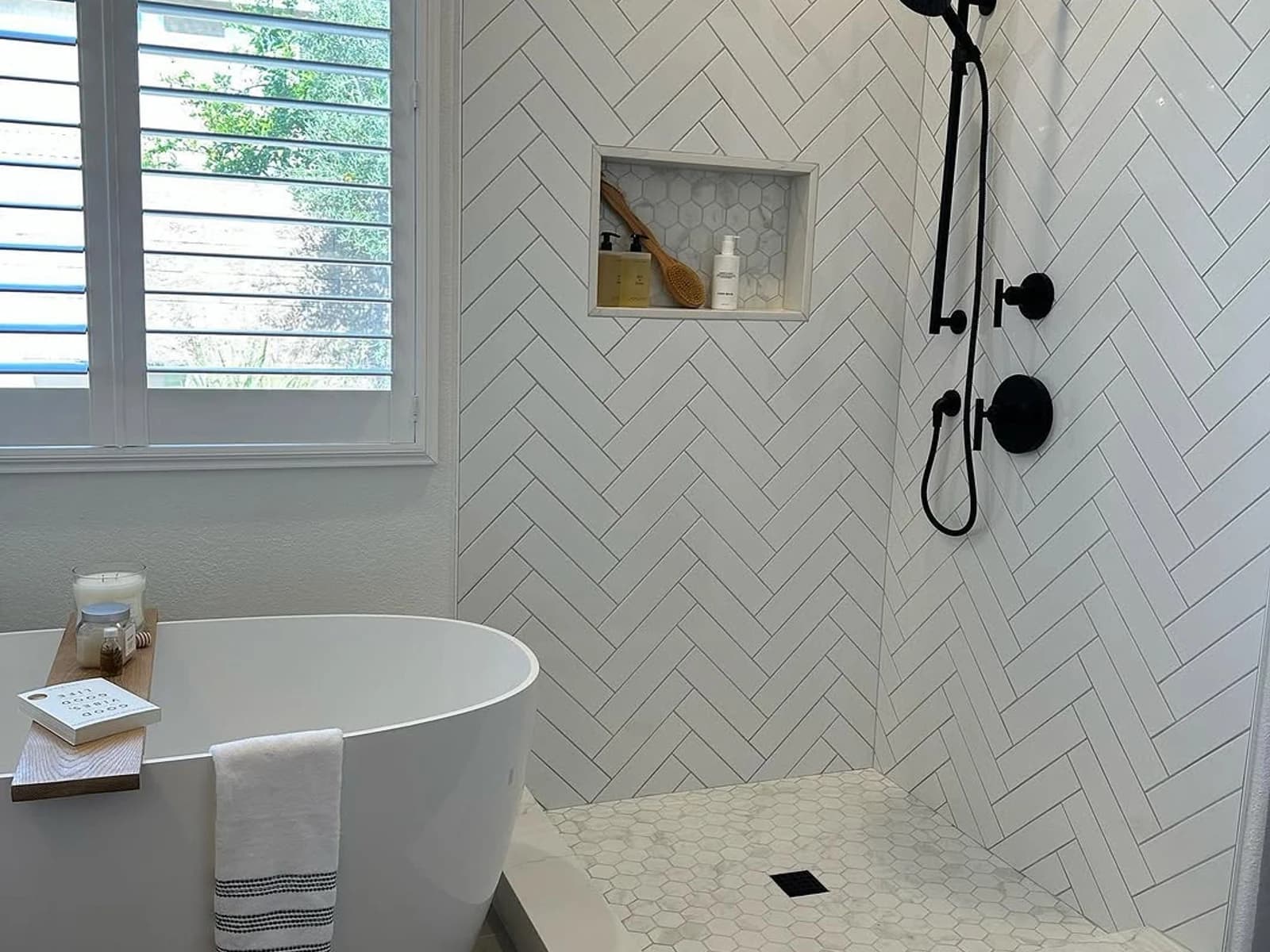 Bright bathroom with freestanding tub, white herringbone shower wall tile, window shutters, and black fixtures