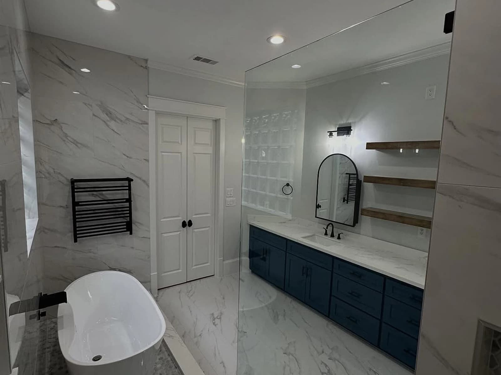 Finished bathroom with a freestanding tub, glass shower, blue vanity, floating shelves, and marble-look tile