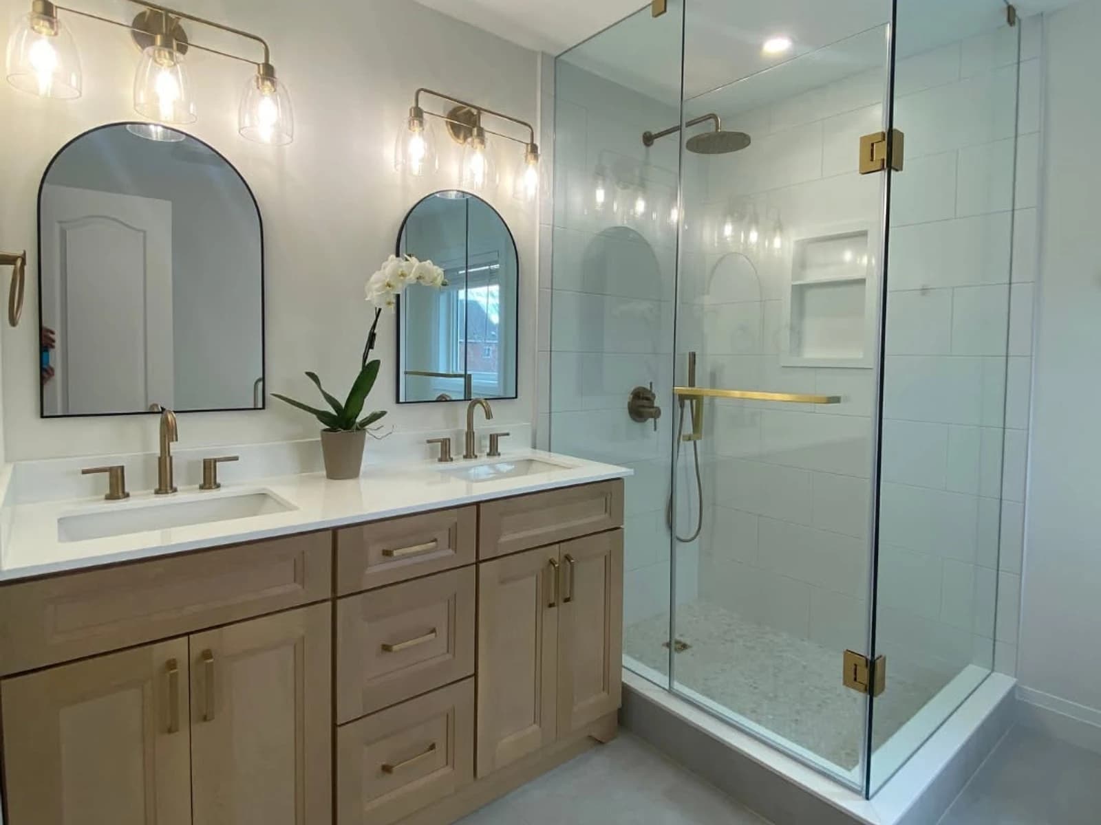 Bathroom remodel with a wood vanity, twin mirrors, glass shower, brass hardware, and pale floor tile