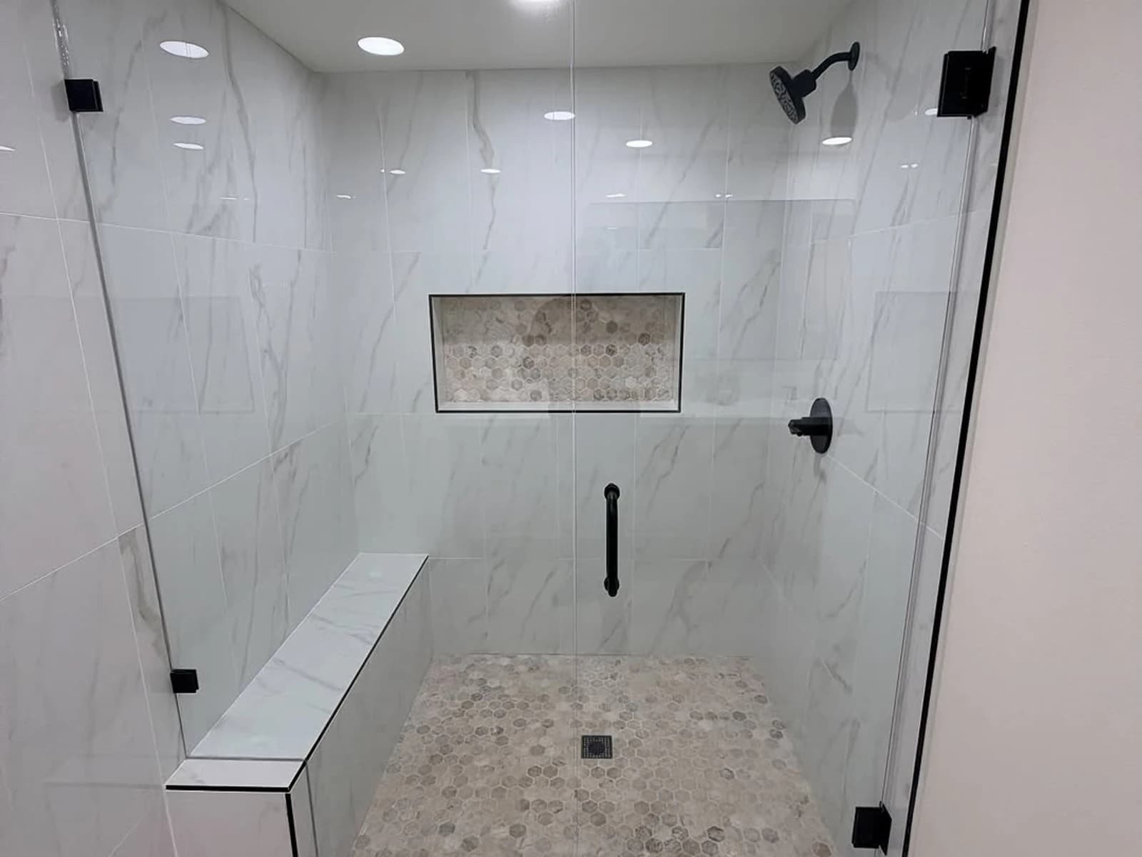 Glass-enclosed shower with marble-look wall tile, black hardware, a bench, and a recessed niche