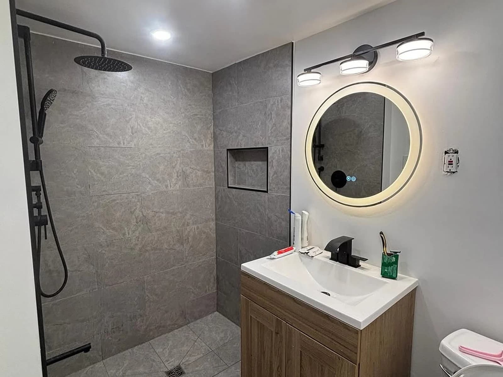 Bathroom with a gray tile walk-in shower, wood vanity, round mirror, and black fixtures