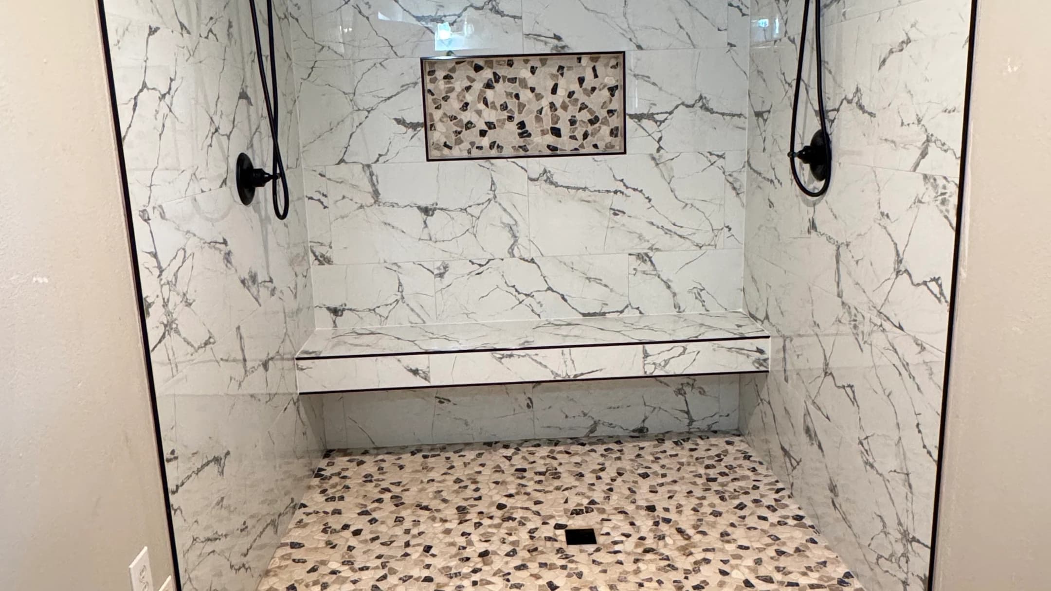 Custom shower with veined wall tile, full-width bench, mosaic niche, dual black fixtures, and mixed-tone floor in Marshall TX